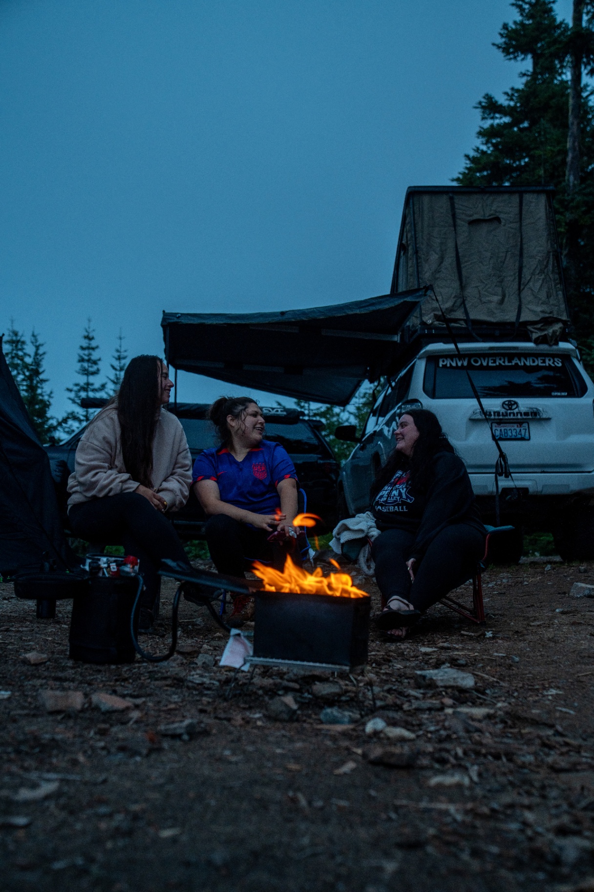Friends gathered around a propane fire pit at dusk with rooftop tent